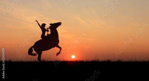 Cowgirl riding a horse, rearing up at sunset. Stallion standing on hind legs at horizon line with setting sun.