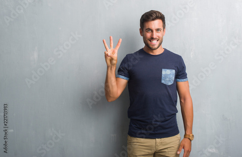 Handsome young man over grey grunge wall showing and pointing up with fingers number three while smiling confident and happy.