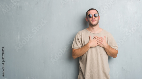 Young caucasian man over grey grunge wall wearing sunglasses smiling with hands on chest with closed eyes and grateful gesture on face. Health concept.