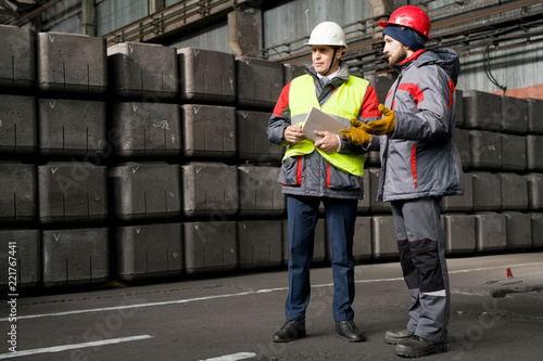 Full length portrait of mature foreman wearing hardhat giving instructions to worker while standing by concrete blocks in workshop, copy space