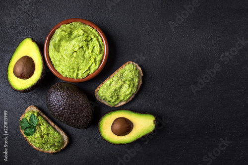 Traditional Mexican Dip Sauce Guacamole in a bowl with bread toasts, whole and cut half avocado on dark background. Top view. Copy space.