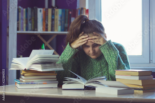 sad woman in a green sweater at a table in the middle of a stack of books and study materials, a blurred bookcase and a window for the background