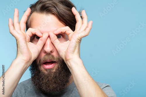 funny ludicrous joyful comic playful man pretending to look through binoculars made of hands. portrait of a young bearded guy on blue background. emotion facial expression concept