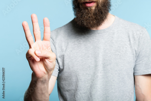 bearded man showing number three with his hand. cropped shot of a male torso on blue background. casual hipster in grey t-shirt counting gesture.