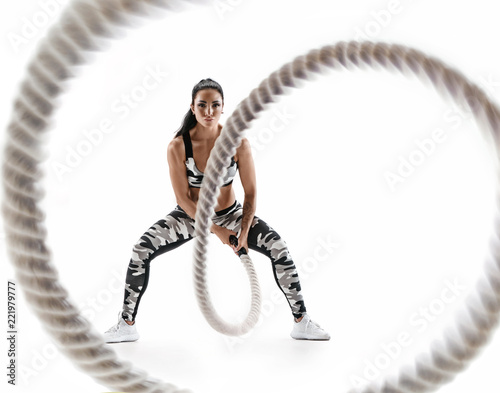 Woman doing exercises with battle rope. Photo of muscular model in military sportswear isolated on white background. Strength and motivation