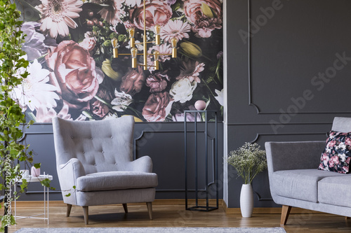 Grey armchair against flowers wallpaper in dark living room interior with sofa and plant. Real photo
