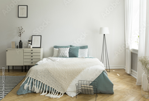 A big comfortable bed with pale sage green and white linen, pillows and blanket in a woman's bright bedroom interior with windows. Real photo.