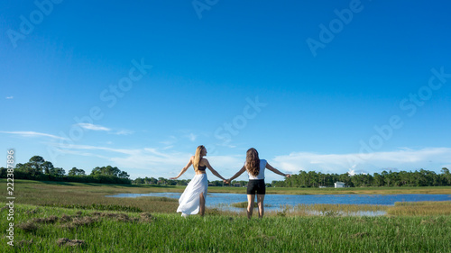 Two women friend blonde & brunette with long hair holding hands in a field next to a lake in florida