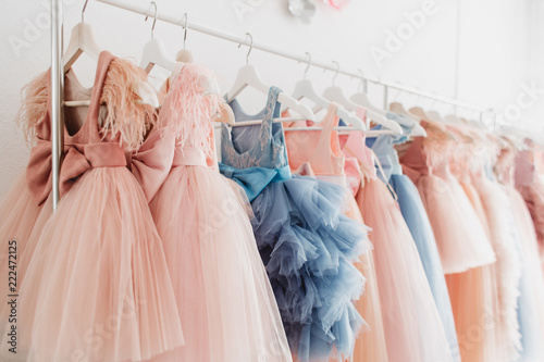 Beautiful dressy lush pink and blue dresses for girls on hangers at the background of white wall. Kids dresses with feathers for prom and holiday.