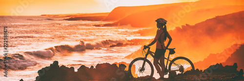 Sport biking cyclist looking at ocean beach coastline during cycling trail race trip. Silhouette of woman athlete training mountain bike outdoors at sunset, banner panorama.