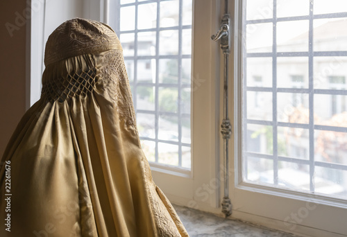 Indoor head and shoulder shot of a person wearing a gold-coloured Burqa looking through a grilled window to the outside