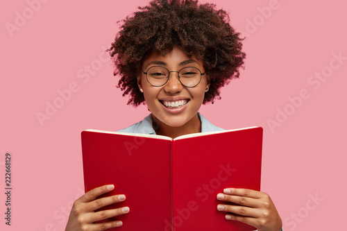 Indoor shot of glad Afro American bookworm holds interesting book in front, has broad smile, happy to finish reading detective story, spends free time at home, stands over pink studio background