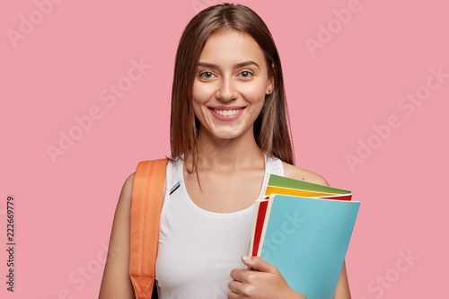 Horizontal shot of pleasant looking cheerful European girl with dark straight hair, dressed in casual white t shirt, carries rucksack and textbooks, isolated over pink background. Emotions, learning