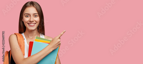 Studio shot of positive Caucasian woman with shining smile, dark straight hair, poins at upper right corner, shows free space for your advertisement, ready for school, isolated over pink background