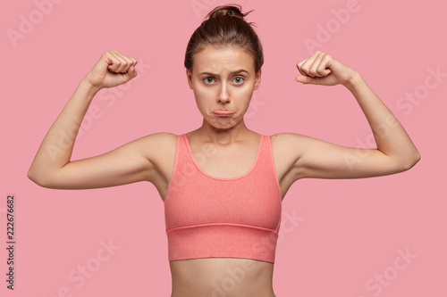 Self detrermined fit woman with sporty body, shows muscles, wears casual top, purses lips, ready for workout and physical training, isolated over pink background. People and motivation concept