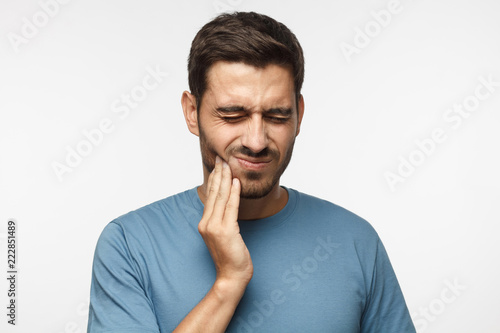 Tooth ache concept. Indoor shot of young male feeling pain, holding his cheek with hand, suffering from bad toothache, looking at camera with painful expression