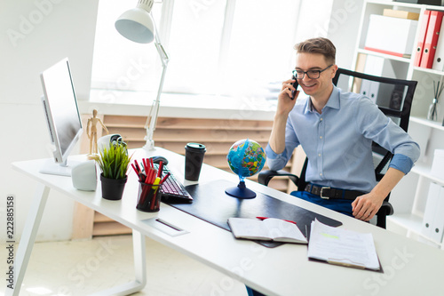 A young man sits in the office at a computer desk, talking on the phone and looking at the globe.