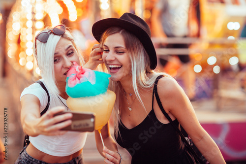 Happy female friends in amusement park eating cotton candy and taking selfie.Two young women enjoying a day at amusement park. 