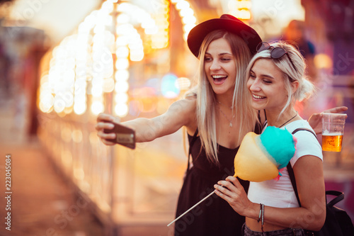 Happy female friends in amusement park eating cotton candy and taking selfie.Two young women enjoying a day at amusement park. 