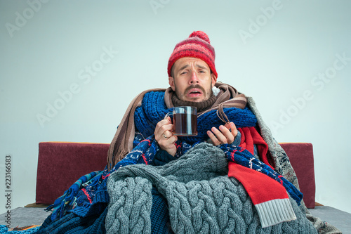 Bearded sick man with flue sitting on sofa at home or studio with cup of tea covered with knitted warm clothes. Illness, influenza concept. Relaxation at Home. Healthcare Concepts.