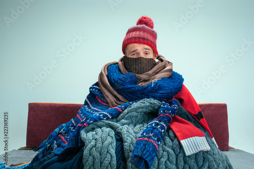 Bearded sick man with flue sitting on sofa at home or studio covered with knitted warm clothes. Illness, influenza, pain concept. Relaxation at Home. Healthcare Concepts.