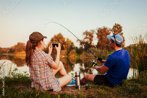 Young couple fishing and drinking tea on river at sunset. Woman filming her boyfriend catching fish. People having fun