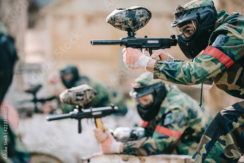 selective focus of paintball player in protective mask aiming with marker gun and his teammate behind outdoors