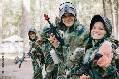 smiling young male paintballer embracing female teammate in camouflage with paintball gun outdoors