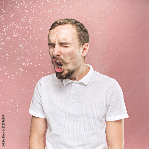 Young funny handsome man with beard and mustache sneezing with spray and small drops, studio portrait on pink background. Comic, caricature, humor. illness, infection, ache. Health concept