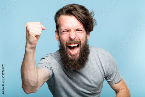 victory success and achievement. excited thrilled agitated guy making a win gesture. hipster man portrait on blue background. emotional reaction and facial expression concept.