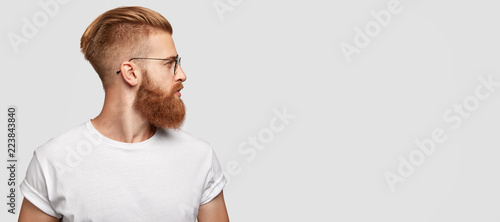 Sideways shot of brutal man with ginger thick beard, trendy hairstyle, looks thoughtfully aside, wears round spectacles and casual t shirt, stands over blank studio background with free space
