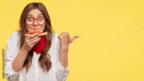 Lunch time concept. Attractive cheerful woman eats delicious Italian pizza, surprised with wonderful taste, indicates with thumb aside, shows where pizzeria situated, suggests to visit and have snack