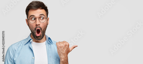 Astonished impressed speechless guy with jaw dropped, points right on copy space with thumb, shows something terrible, wears round spectacles and fashionable shirt. Omg and advertisement concept