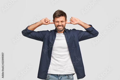 Troubled middle aged man pluggs ears with displeased expression, doesnt want to hear advice from friends, ignores annoying sound, clenches teeth, dressed in fashionable shirt, isolated on white