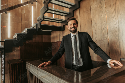 Welcome to our hotel. Portrait of cheerful receptionist looking at camera while standing behind wooden hostess desk