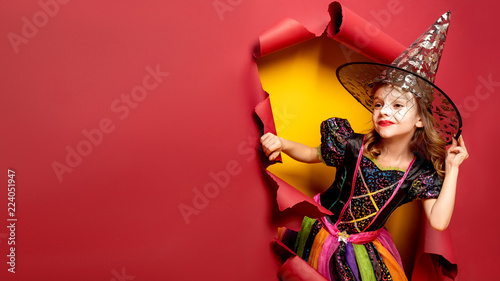 Happy Halloween. Laughing funny child girl in a witch costume of halloween looking and smiling through a hole of red, yellow paper background. Copyspace. 16 to 9 size