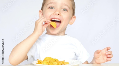 A child with pleasure eating potato chips sitting at a table on a white background. The boy is eating chips and smiling.