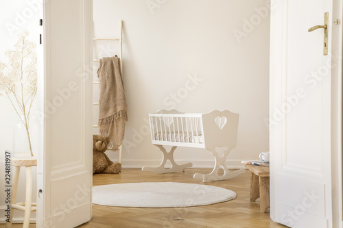 Real photo of white cradle with heart standing in Scandi kid room interior with teddy bear, herringbone parquet and empty wall with place for your poster