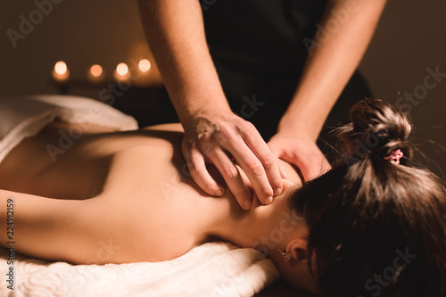 Men's hands make a therapeutic neck massage for a girl lying on a massage couch in a massage spa with dark lighting. Close-up. Dark Key