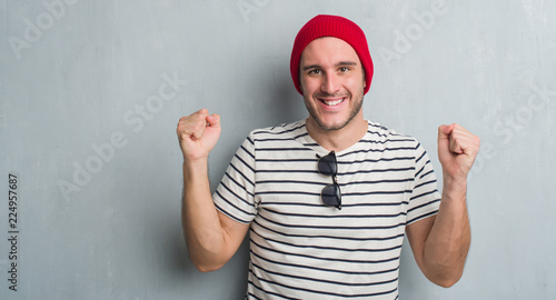 Young caucasian man over grey grunge wall wearing wool cap screaming proud and celebrating victory and success very excited, cheering emotion