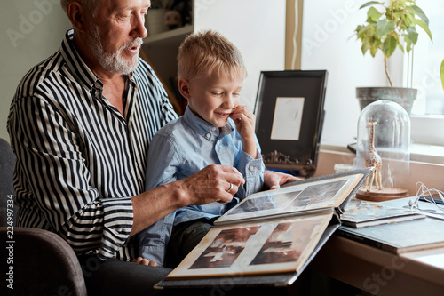 senior man and little boy holding and looking at family photo album in living room