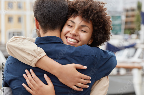 Farewell concept. Delighted happy smiling African American woman says goodbye to boyfriend who sails for long time, gives warm hug, pose together against harbour background. Truthful feelings