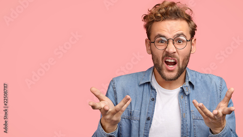 Annoyed young Caucasian guy gestures with hands angrily, opens mouth and frowns face in discontent, poses against pink background with copy space for your advertisement or promotion, asks what to do