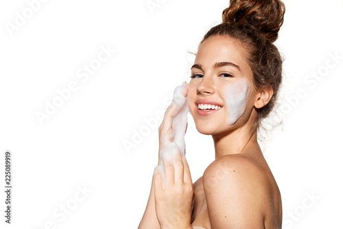 Portrait of young pretty girl with healthy skin on white background. Woman smiling and applying cosmetic foam on face. Beauty skincare concept.