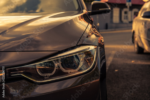 grille and headlight of a brown car in a parking lot against the background of a blurred old house with morning sunbeams