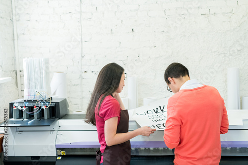 Rear view of busy printing specialists examining banner with text and standing at large format printer in office