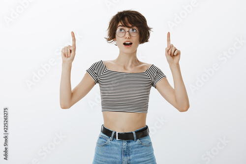 Wow look there. Portrait of curious and impressed intrigued woman in round glasses and cropped top, raising index fingers and pointing up while looking at upper left corner with amazement