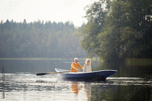 Happy senior couple in casual clothing sitting on boat and enjoying romantic date on lake, elderly man rowing oars