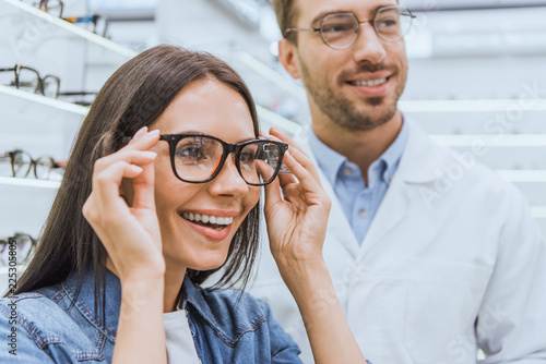 portrait of smiling woman choosing eyeglasses while male optometrist standing near in optics