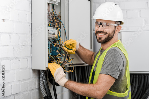 smiling handsome electrician repairing electrical box with pliers in corridor and looking at camera